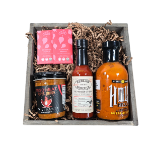 A rustic wooden crate filled with spicy gourmet items including Hot Reaper Miso, a Scorpion Chocolate Bar, Blonde Beard's hot sauce, and Redlaw Trinidad Scorpion Hot Sauce