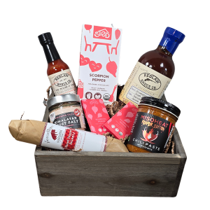 A wooden crate filled with a selection of spicy foods including ghost pepper salami, scorpion pepper chocolate, ghost salt, reaper miso, blueberry ghost barbecue sauce, and reaper hot sauce, all set against a rustic wooden background.