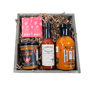 A rustic wooden crate filled with spicy gourmet items including Hot Reaper Miso, a Scorpion Chocolate Bar, Blonde Beard's hot sauce, and Redlaw Trinidad Scorpion Hot Sauce